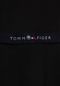 Czarna tkanina z pasem w talii, na którym znajduje się napis "TOMMY HILFIGER" w białym kolorze, ozdobiony małym czerwonym i białym logo oraz czarnym guzikiem.