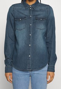 Dark blue denim shirt with long sleeves, button front, two chest pockets, and a classic collar. Features a curved hem and contrasting stitching.
