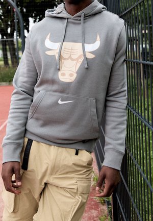 Hombre con sudadera gris con capucha con un logo estilizado de toro y el símbolo de Nike, pantalones cargo beige, de pie junto a una cerca metálica en una cancha deportiva.