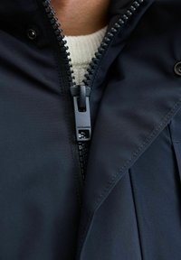Navyblauer Jacke mit einem Frontreißverschluss, strukturiertem Stoff und einem cremefarbenen Pullover darunter. Mit einem Logo am Reißverschlussanhänger.