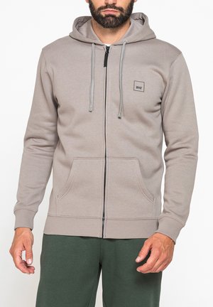 Uomo che indossa una felpa beige con cappuccio e zip, con coulisse e tasche anteriori, abbinata a pantaloni verde scuro, su sfondo bianco.