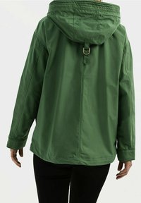 Personne portant une veste à capuche verte ample avec un détail en anneau métallique à l'arrière, associée à un pantalon noir, debout de dos.