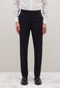 Klassieke zwarte pantalon met een getailleerde pasvorm, voorzien van een vlakke voorkant, riemlussen en een gladde textuur. Gecombineerd met zwarte veterschoenen.