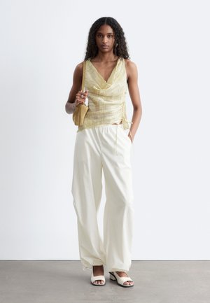 Žena oblečená v zlatej tielkovej blúzke, bielych nohaviciach s širokými nohavicami, bielych otvorených opätkoch a nesie malú hnedú kabelku na ramene, stojaca pri jednofarebnej stene.