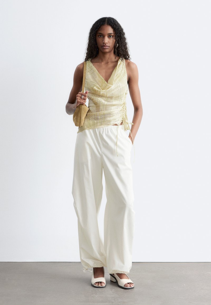 Femme portant un top doré sans manches, un pantalon blanc à jambes larges, des talons ouverts blancs et portant un petit sac à bandoulière beige, debout contre un mur uni.