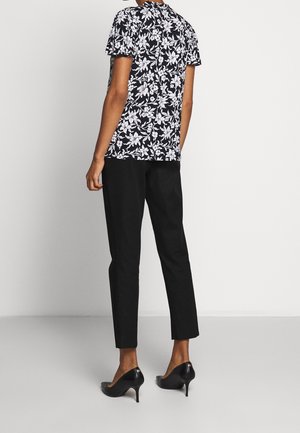 Personne debout de dos portant un haut à manches courtes à fleurs noir et blanc, un pantalon noir à la cheville et des chaussures noires à talons hauts.