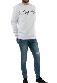Witte sweatshirt met zwarte tekst "Project x Paris", geribbelde manchetten en onderkant, gecombineerd met lichtblauwe versleten jeans en zwarte sneakers.