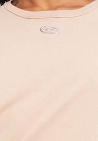 Gaiši ādas krāsas T-krekls no gludas auduma. Raksturo apaļu kakla izgriezumu un nelielu sudraba logo krūtīs. Mīksta tekstūra ar minimālām šuvēm.