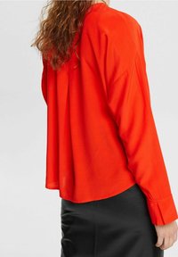 Blusa rossa a maniche lunghe con vestibilità comoda, caratterizzata da un colletto arrotondato e maniche con polsini, abbinata a una gonna nera. Tessuto dalla consistenza liscia.