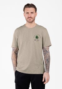 Hombre con cabello corto y tatuajes en ambos brazos, vistiendo una camiseta beige con un pequeño gráfico de una palmera verde en el pecho y pantalones negros.