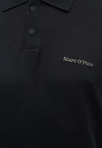 Μαύρο πόλο με υφή, δύο μαύρα κουμπιά και το «Marc O’Polo» κεντημένο με λευκό στο στήθος.