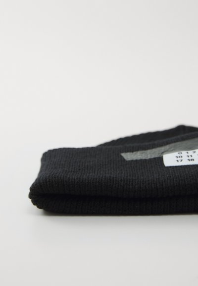Fållad svart stickad mössa med en grå rand och en vit etikett som visar numeriska designdetaljer på en vit yta.