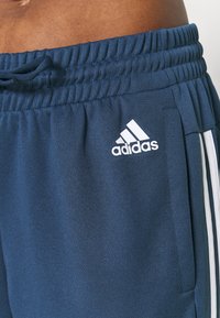 Gros plan sur un pantalon de sport Adidas bleu marine avec logo blanc et bandes latérales, doté d'une taille élastique et d'un cordon de serrage.