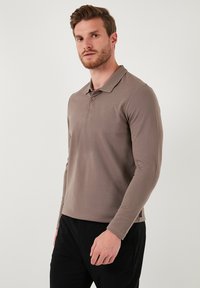 Polo de manga larga marrón hecho de un tejido suave con cuello de canalé y tapeta de dos botones, combinado con pantalones negros.