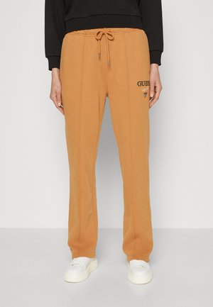 Persoon draagt beige trekkoordbroek met verticale naden en "GUESS"-logo op de dij, gecombineerd met witte sneakers en zwarte top.