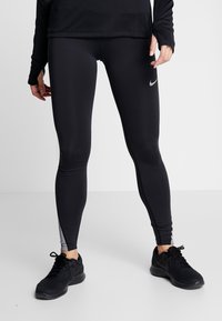 Persoon in zwarte Nike-legging en zwarte hardloopschoenen staat op een lichtgrijze vloer tegen een witte muur.