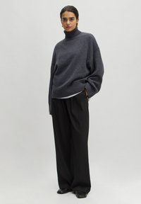 Flauschiger grauer Strickrollkragenpullover mit geripptem Saum, kombiniert mit lockeren schwarzen Hosen und schwarzen Schuhen, die eine lässige Silhouette schaffen.