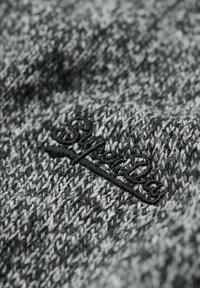 Szary i czarny dzianinowy materiał z teksturowanym wzorem. Zawiera czarne haftowane logo lub detal wzoru na powierzchni.