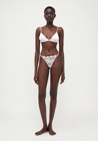 Valkoinen alusvaatesetti, jossa on kolmionmuotoinen rintaliivit ohueilla olkaimilla ja siihen sopivat pitsiset stringit. Kangas on sileä, koristeltu kukallisilla pitsidetaljeilla.