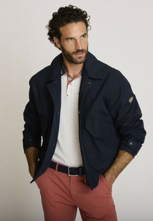 Homme aux cheveux bouclés portant une veste bleu marine, un polo blanc et un pantalon corail, debout les mains dans les poches, regardant sur le côté.