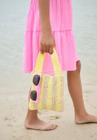 Otrok v roza obleki drži rumeno progasto tkan torbo z roza sončnimi očali, stoji boso na peščeni plaži blizu vode.