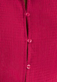 Camisa de botón en fucsia con una tela texturada y arrugada. Presenta tres botones brillantes rosa en la parte delantera y un acabado suave.