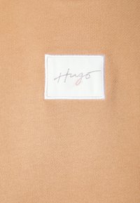Tela beige tejida con una etiqueta blanca rectangular que presenta la palabra "Hug" en letras cursivas grises en la esquina superior izquierda.