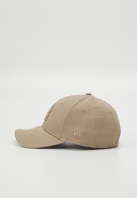 Casquette de baseball beige avec une couronne arrondie, une visière courbée, un logo brodé à l'avant et sur le côté, ainsi que des œillets de ventilation.