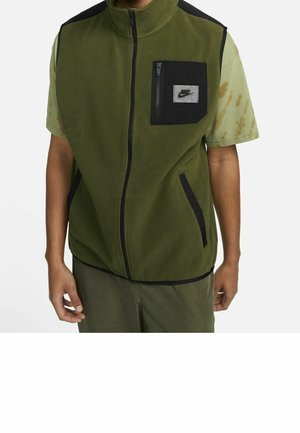 Homme portant un gilet polaire vert avec bordure noire et poche poitrine zippée par-dessus une chemise verte à manches courtes avec des taches marron.