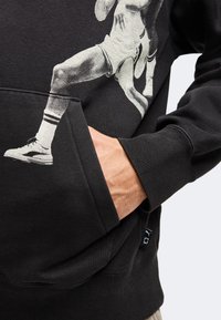 Main dans la poche d’un sweat-shirt noir avec un graphisme blanc montrant les jambes d’un joueur de basket portant un short, des chaussettes et des baskets.