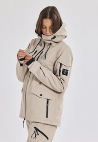 Beige wetterfeste Jacke mit hohem Kragen, verstellbarem Kapuze und schwarzen Reißverschlüssen. Verfügt über Markenlogos und Daumenlöcher an den Ärmelabschlüssen für zusätzliche Wärme.