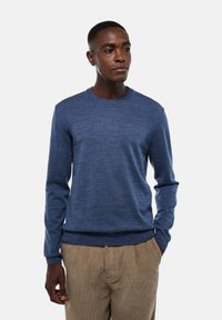 Homme portant un pull à manches longues bleu et un pantalon en velours côtelé beige, se tenant avec une main dans la poche sur un fond uni.