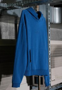 Niebieska bluza z kapturem wisząca na metalowym stojaku w przemysłowym otoczeniu, z kanałami wentylacyjnymi i metalowymi kratami w tle.
