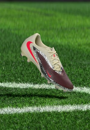 Nogometni čevelj Nike v bež in bordo barvi z rdečimi logotipi Swoosh, vzorčastimi poudarki in vezalkami, ki lebdi nad označenim zelenim travnjakom.