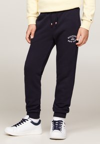 Person in marineblauen Jogginghosen mit weißem "Hilfiger 1985 New York" Logo und weißen Sneakers mit marineblauen Schnürsenkeln, vor beigem Hintergrund stehend.