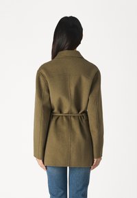 Abrigo verde olivo con cinturón, cuello estructurado y mangas largas. El tejido parece texturizado y cálido, con un corte recto en el dobladillo.