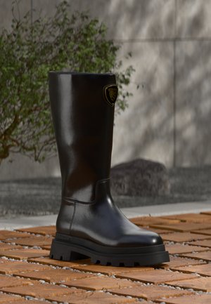 Stivale nero in pelle fino al ginocchio con suola spessa in gomma e battistrada testurizzato, posizionato all'aperto su pavimento in legno e pietra con piante sullo sfondo.