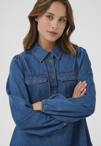 Tumši zila džinsa krekls ar apkakles kaklu, divām krūšu kabatām, pogu aizdari un savirknētām piedurknēm, nodrošinot komfortablu piegriezumu.