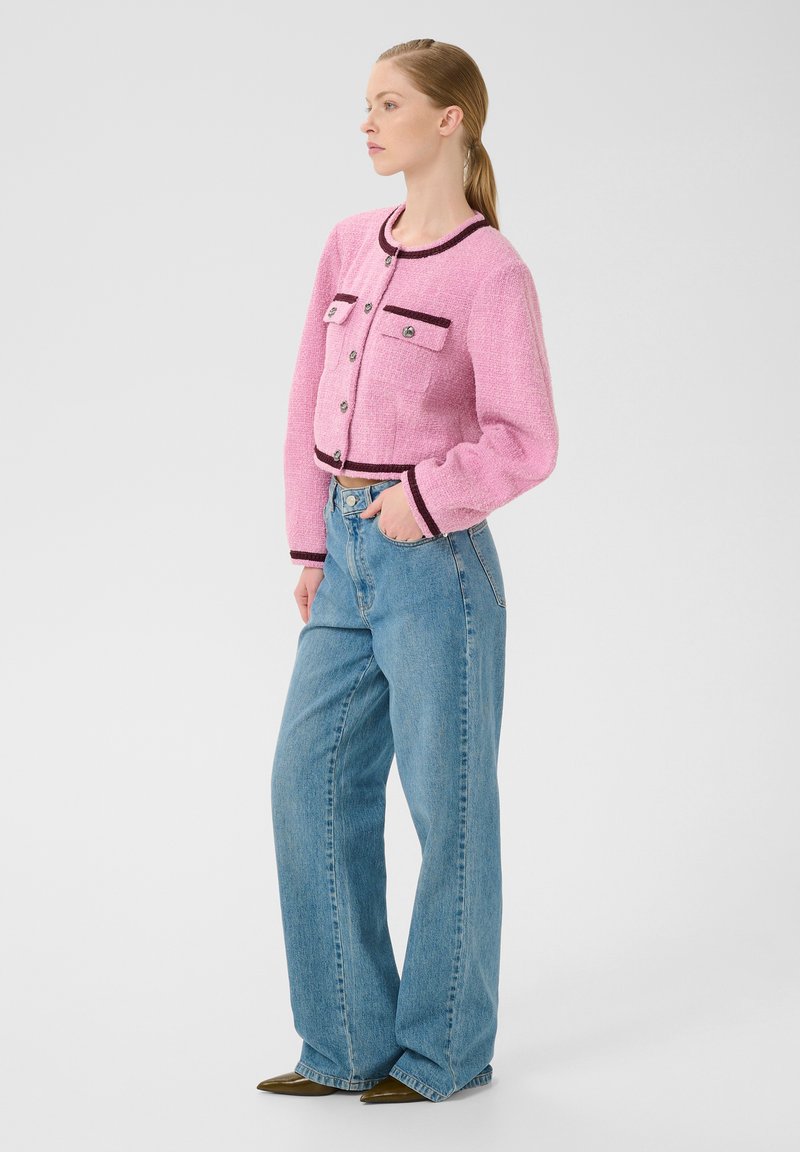 Rosa tweed kort jacka med svarta detaljer och knappar, med två framfickor och långa ärmar, i kombination med ljusblå vida jeans.