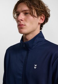 Giacca zip-up navy in tessuto morbido con colletto alto, caratterizzata da un design del logo bianco sul lato sinistro del petto.