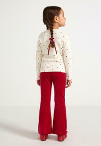 Langærmet cremefarvet top med print af rødt hjerte, med detalje af sløjfe bagpå, kombineret med højtaljede røde velourbukser med vidde.