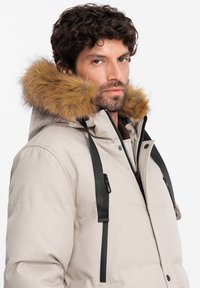 Parka imbottita beige chiaro con rifiniture in pelle ecologica sul cappuccio, dettagli in metallo nero e cinghie regolabili sulle spalle per una vestibilità sicura.