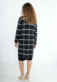 Cardigan à carreaux noir et blanc avec manches longues, design mi-long et texture douce, porté par-dessus une jupe noire et des talons rose clair.