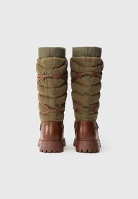 Φουσκωτές μπότες μέχρι το γόνατο σε ελιά πράσινο με καφέ λαστιχένιες σόλες, με λουράκια στο πλάι και ριμπ πλεκτές μανσέτες για υφή στο φινίρισμα.