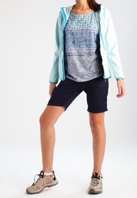 Ljusblå zip-up jacka över en mönstrad ljusblå och grå topp, tillsammans med marinblå shorts och beige vandrarskor med röda accenter.