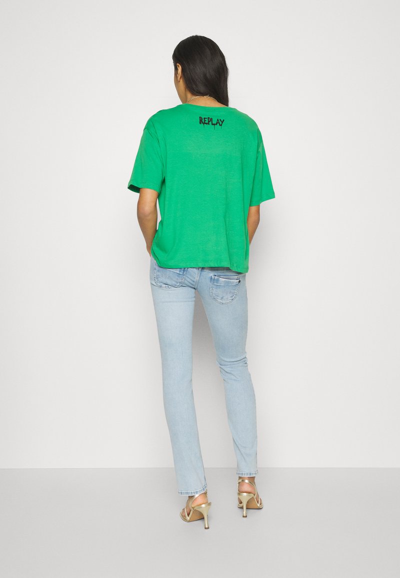 Grünes Baumwoll-T-Shirt mit kurzen Ärmeln und lässigem Schnitt, mit schwarzem "REPLAY"-Text auf der Rückseite. Kombiniert mit hellblauen Jeans.