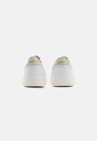 Zapatillas blancas con parte superior de cuero liso, suaves detalles en gris y suela de goma. Logotipo "Reebok" en amarillo en el talón.