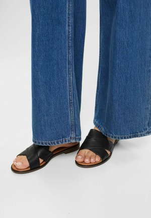 Black leather slide sandals with crisscross strap design, low heel, and open toe, paired with wide blue denim pants.