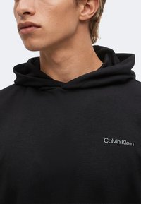 Νεαρός άνδρας που φοράει μαύρο φούτερ Calvin Klein, ορατός από το λαιμό μέχρι το πάνω μέρος του στήθους, κοιτάζοντας αριστερά μπροστά σε ένα μονόχρωμο φόντο.