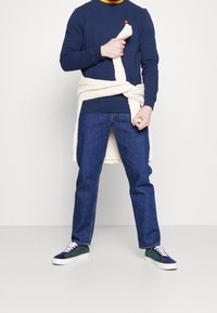 Navy sweatshirt with a round neck, a small red detail, paired with blue jeans and green sneakers with white soles and multicolored laces.